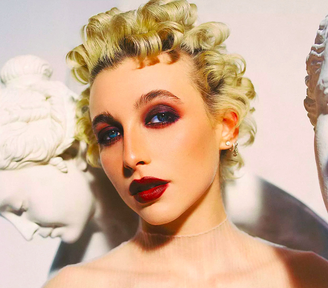 Emma Chamberlain with curled blonde hair and a deep red lip and eye posing among statues.