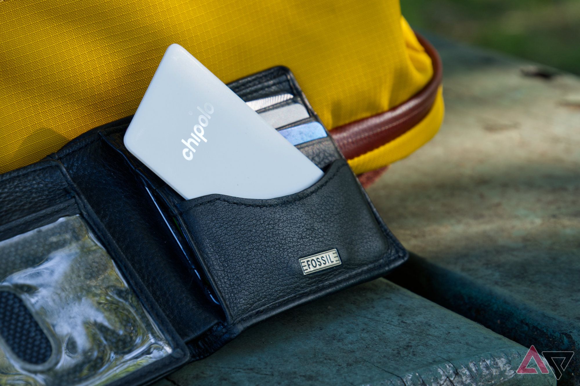 Chipolo CARD Point sticking partially out of an open leather wallet resting against a bright yellow bag on a picnic table